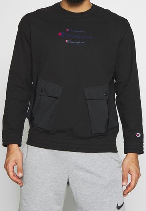 Czarna bluza Champion z trzema logo Champion na piersi i dużymi kieszeniami na guziki, noszona z jasnoszarymi szortami Nike.