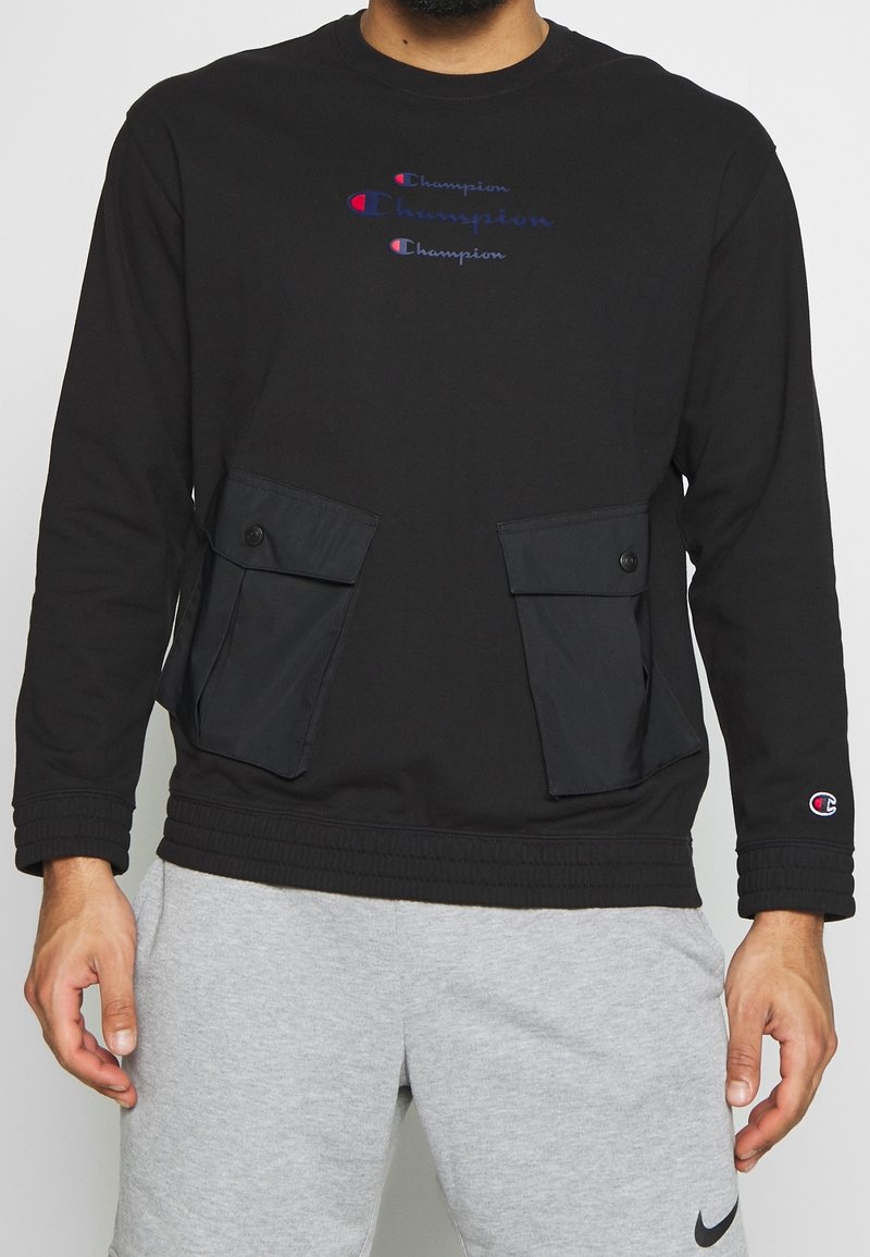 Sweat-shirt noir Champion avec trois logos Champion sur la poitrine et grandes poches boutonnées, porté avec un short Nike gris clair.
