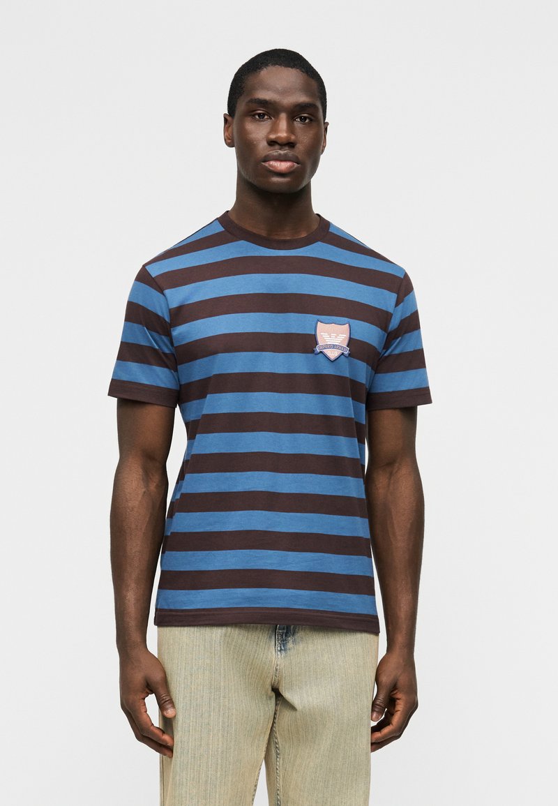 Άνδρας που φοράει μπλε και καφέ ριγέ μπλουζάκι με μικρό έμβλημα στο στήθος, συνδυασμένο με τζιν ανοιχτού χρώματος, στέκεται μπροστά σε απλό φόντο.