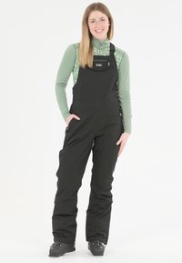 Schwarze Schneehosen mit verstellbaren Trägern und Reißverschlusstaschen, kombiniert mit einem grünen Langarmshirt, das ein dezentes Muster aufweist.