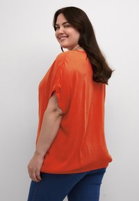 Blouse orange vif à manches courtes, coupe décontractée et texture lisse, associée à un jean bleu. Design simple, sans motifs ni accents visibles.