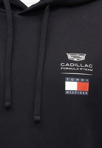 Sweat à capuche noir avec le logo de l'équipe Cadillac Formula 1 au-dessus du logo du drapeau Tommy Hilfiger sur la poitrine, avec des cordons au niveau de l'encolure.