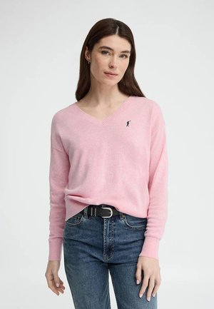 Femme aux longs cheveux bruns portant un pull rose clair à col en V et un jean bleu, debout devant un fond clair uni.