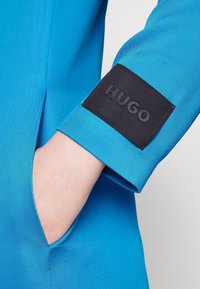 Jasnoniebieska tekstylna kurtka o fakturowanej powierzchni, z czarną metką "HUGO" na mankiecie. Posiada boczną kieszeń.