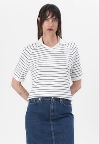 Hvit kortermet polo skjorte med marineblå striper, med v-hals og et logoemblem på brystet. Kombinert med et mørkt denimskjørt.