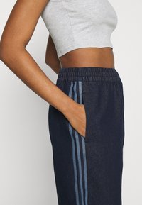 Person wearing a gray cropped tank top and dark blue track pants with light blue side stripes, hand casually placed in pocket.