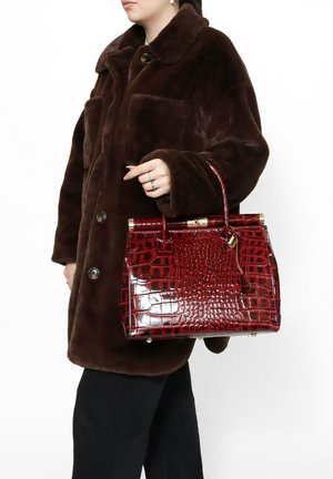 Rode handtas met krokodillenembossing, gestructureerde vorm, gouden hardware en een bovenste handvat, gecombineerd met een bruine bontjas.