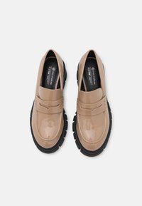 Mocassins en cuir verni beige avec une sangle texturée, semelle en caoutchouc noir, bout rond et détails cousus.