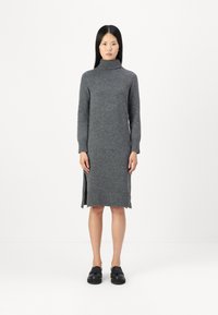 Pilkos vilnos turtleneck suknelė ilgomis rankovėmis, su raukiniais ant rankovių ir apykaklės, bei šoninėmis plyšėmis. Modelis vilki juodus batelius, stovėdamas prieš baltą foną.
