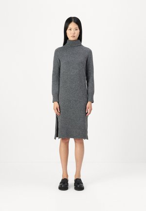 Robe en laine grise à col roulé avec des manches longues, des poignets et un ourlet côtelés, et des fentes sur les côtés. Le modèle porte des chaussures noires, se tenant contre un fond blanc.