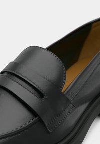 Μαύρο δερμάτινο μοκασίνι με λεία υφή, που διαθέτει ανάγλυφη λεπτομέρεια λουριού μπροστά και εσωτερικό σε ανοιχτό καφέ χρώμα.