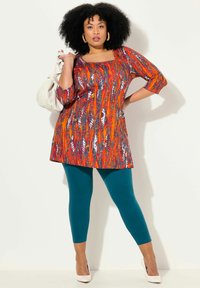 Túnica colorida con escote cuadrado en un patrón abstracto naranja y azul, combinada con leggings teal y tacones blancos. Diseño ajustado y de longitud a la rodilla.