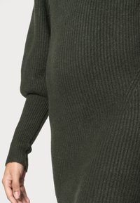 Pull vert tricoté avec une texture côtelée, manches longues et coupe ample, une main visible tendue vers le vêtement.