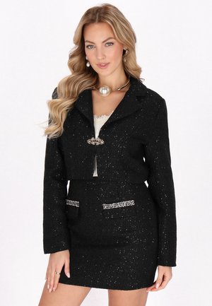 Jachetă din țesătură tweed neagră, cu un design scurt, accente din metal argintiu și buzunare, purtată peste un top deschis la culoare, asortată cu o fustă similară.