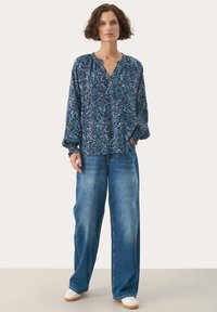 Blouse à manches longues bleu à motifs avec une coupe relaxe, associée à un jeans en denim bleu clair à coupe ample et des baskets blanches avec des semelles beiges.
