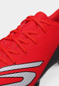 Κοντινό πλάνο κόκκινου αθλητικού παπουτσιού με υφή στην επιφάνεια, κόκκινα κορδόνια και μεγάλο ασημί και μαύρο στυλιζαρισμένο λογότυπο στο πλάι.