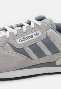 Sivé semišové tenisky s priehľadnými akcentmi, bielymi šnúrkami a tromi sivými pruhmi. Logo Adidas na bielom štítku jazyka. Gumová podrážka.