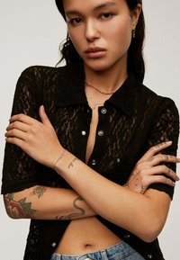 Camisa negra de encaje con botones y mangas cortas, botones plateados y cuello abierto. El modelo lleva collares en capas y joyería de plata, mostrando tatuajes.