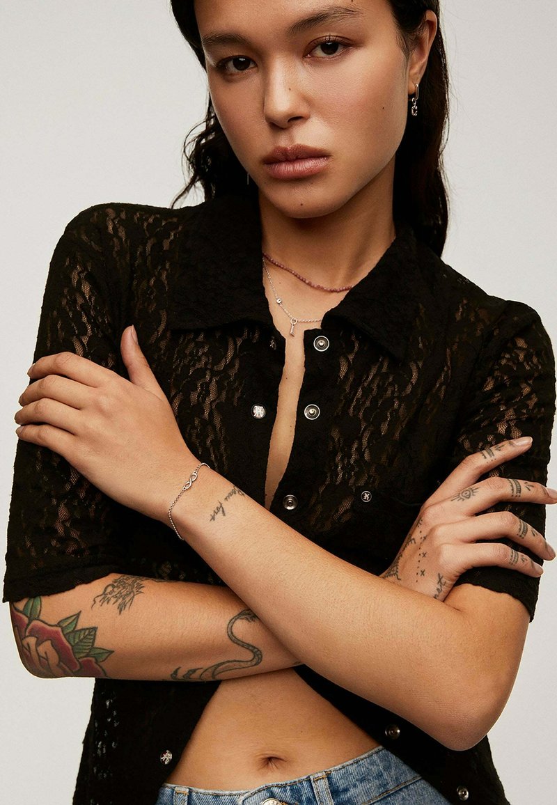 Camisa negra de encaje con botones y mangas cortas, botones plateados y cuello abierto. El modelo lleva collares en capas y joyería de plata, mostrando tatuajes.