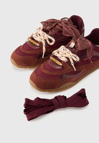 Παπούτσια σνίκερς σε μπορντό χρώμα με υλικά  από σουέτ και δίχτυ, κυρτό σχέδιο και κορδόνια με σχέδιο. Συνοδεύονται από ένα επιπλέον ζευγάρι κορδόνια σε μαρόν.