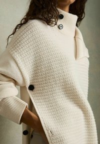 Pull en tricot couleur crème avec un motif texturé, col montant et boutons noirs. Présente des poignets côtelés et une coupe décontractée.