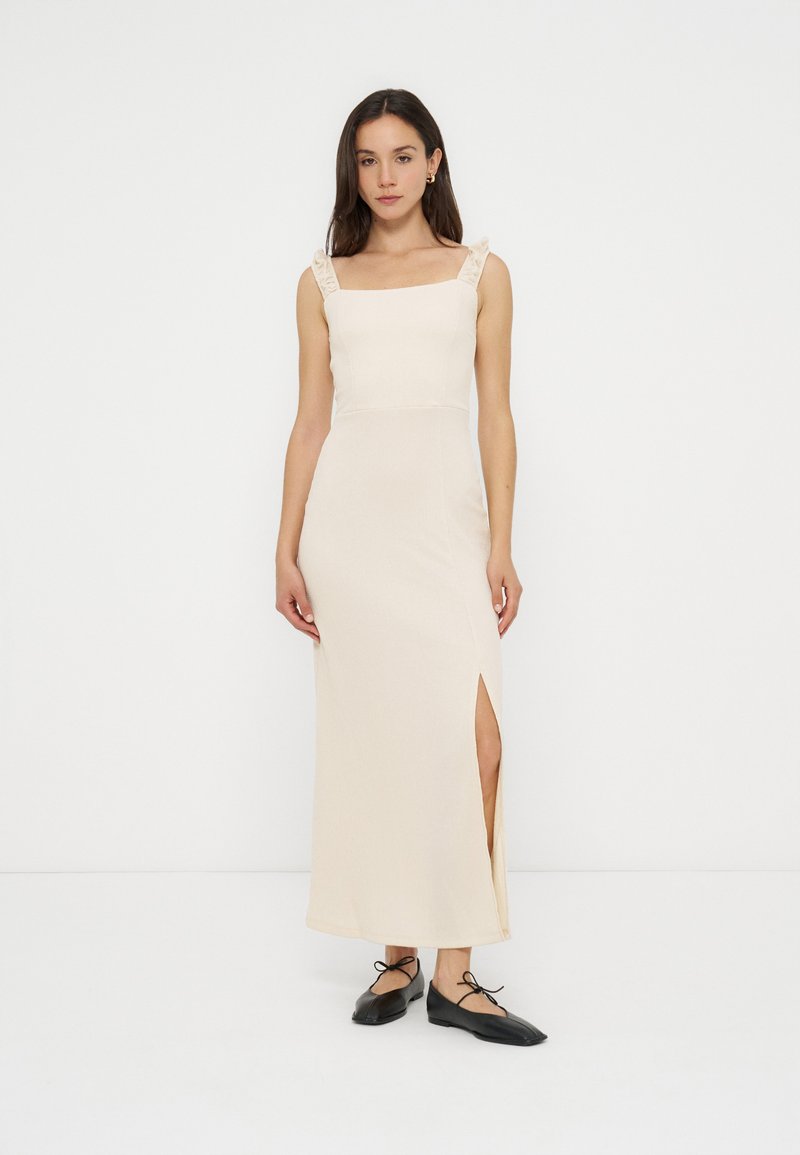 Femme debout portant une robe maxi crème sans manches avec des bretelles à volants et une fente latérale, associée à des ballerines noires à lacets sur fond blanc.