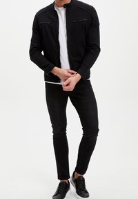 Muž v černé zipové bundě, bílém tričku, černých skinny džínách a černých teniskách s bílou podrážkou na bílém pozadí.