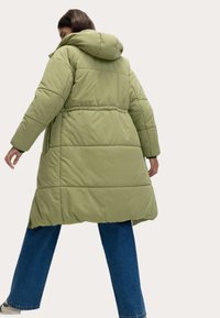 Polstret lang jakke i lysegrøn med hætte, elastisk talje og sidelommer. Kan kombineres med blå vide jeans.