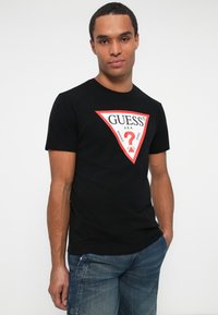 Tricou negru din bumbac cu un logo triunghiular roșu care conține "GUESS", "?" și "WASHED JEANS". Mâneci scurte, guler rotund și un design slim fit.