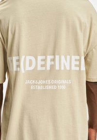 Tan bomull T-skjorte med stor hvit tekst på baksiden: "RE(DEFINED) JACK&JONES ORIGINALS ETABLERT 1990." Korte ermer, avslappet passform.