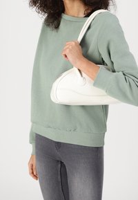 Bolso blanco con forma redondeada, superficie texturizada, dos asas cortas y sutura sutil, sostenido frente a un sudadera verde.