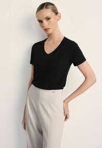 Młoda kobieta z gładko zaczesanymi włosami, ubrana w czarny top z dekoltem w serek i krótkimi rękawami oraz jasnobeżowe spodnie z wysokim stanem, pozująca z jedną ręką za plecami.