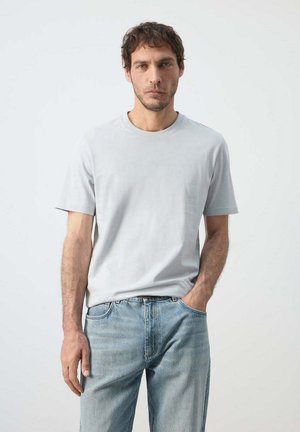 Mann trägt hellgraues kurzärmeliges T-Shirt und ausgewaschene blaue Jeans, steht mit einer Hand in der Tasche vor weißem Hintergrund.
