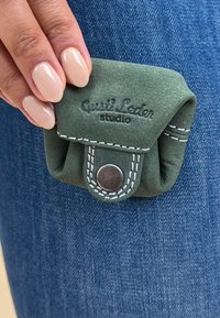 Zelená kožená taška s bílým prošitím, vybavená uzavíráním na cvok a embosovaným logem, držena proti denimové látce.