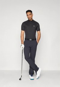 Mustast lühikeste varrukatega golfipluus tõmblukuga, tumesinised püksid, valged golfijalatsid siniste detailidega, hoides golfikeppi ja kandes golfikinnast.