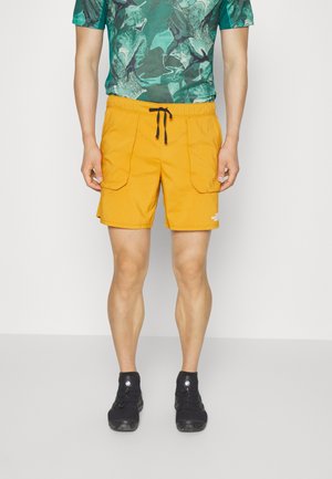 Homme portant un short jaune à cordon avec des poches avant, une chemise verte à motifs et des chaussures de randonnée noires, debout sur un sol gris clair.