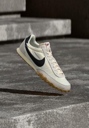 Λευκό και μαύρο αθλητικό παπούτσι Nike με καφέ σόλα, κρεμασμένο πάνω από σκάλες από σκυρόδεμα, φωτισμένο από φυσικό ηλιακό φως.