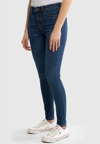 Vaqueros ajustados de denim azul oscuro con un acabado suave. Presentan una cintura alta, cinco bolsillos y detalles de costura sutiles. Se usan con zapatillas blancas.