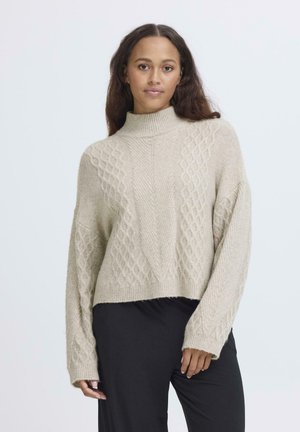 Lys beige strikket sweater med høj hals, der har et struktureret diamantmønster og dropped skuldre. Samlet med sorte bukser.