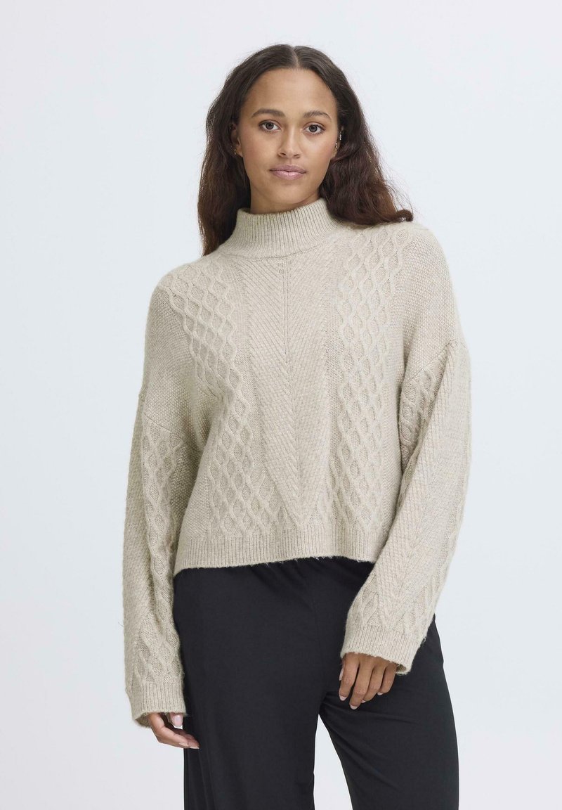 Maglione lavorato a maglia beige chiaro con collo alto, caratterizzato da un motivo a diamante testurizzato e spalle scese. Abbinato a pantaloni neri.