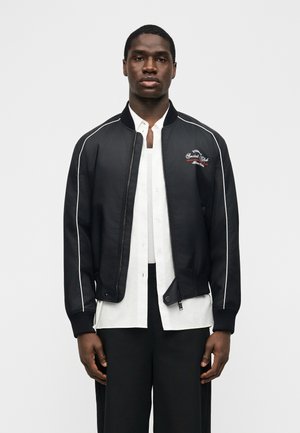 Άνδρας που φορά μαύρο μπουφάν τύπου bomber με λευκές ρίγες και κόκκινο κείμενο πάνω από λευκό πουκάμισο με κουμπιά και μαύρο παντελόνι, στέκεται μπροστά σε απλό φόντο.