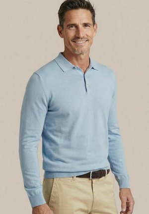 Man glimlacht, draagt een lichtblauw poloshirt met lange mouwen en beige broek met een bruine riem, tegen een effen beige achtergrond.