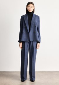 Sieviete, kas valkā zilu dubultpogu kostīmu ar plaša piegriezuma biksēm un melnu augstu apkakli, stāv pret vienkāršu baltu fonu.