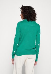 Pull vert en tricot avec un col rond et des accents à pois blancs sur les manches. Tissu texturé et poignets côtelés complètent le design.