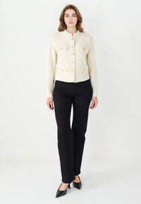 Elfenbeinfarbene Tweedjacke mit kurzem Schnitt, Rundhalsausschnitt und zwei vorderen Taschen. Mit goldfarbenen Knopfverschlüssen und langen Ärmeln.