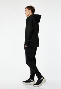 Sort hættetrøje med hætte, reflekterende stribe, kombineret med sorte jogger-bukser og sorte sneakers med strukturerede detaljer. Enkel, sporty design.