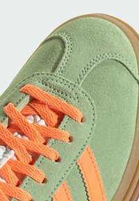 Sneaker verde in suede con lacci e strisce arancioni, caratterizzato da una superficie testurizzata e una suola in gomma. I dettagli includono cuciture a zigzag.