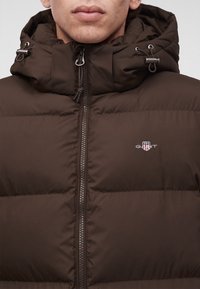 Homme portant une veste matelassée marron avec un col montant et une capuche, zippée, avec un petit logo brodé sur la poitrine.
