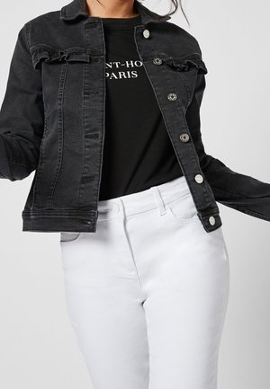 Kvinde iført åben sort denimjakke over sort Saint-Honore Paris t-shirt og hvide bukser med høj talje mod ensfarvet baggrund.