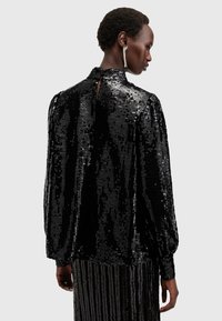 Blusa nera con paillettes, maniche lunghe, scollatura alta e dettaglio cut-out sulla schiena. La texture lucida e il drappeggio fluido migliorano il suo design.